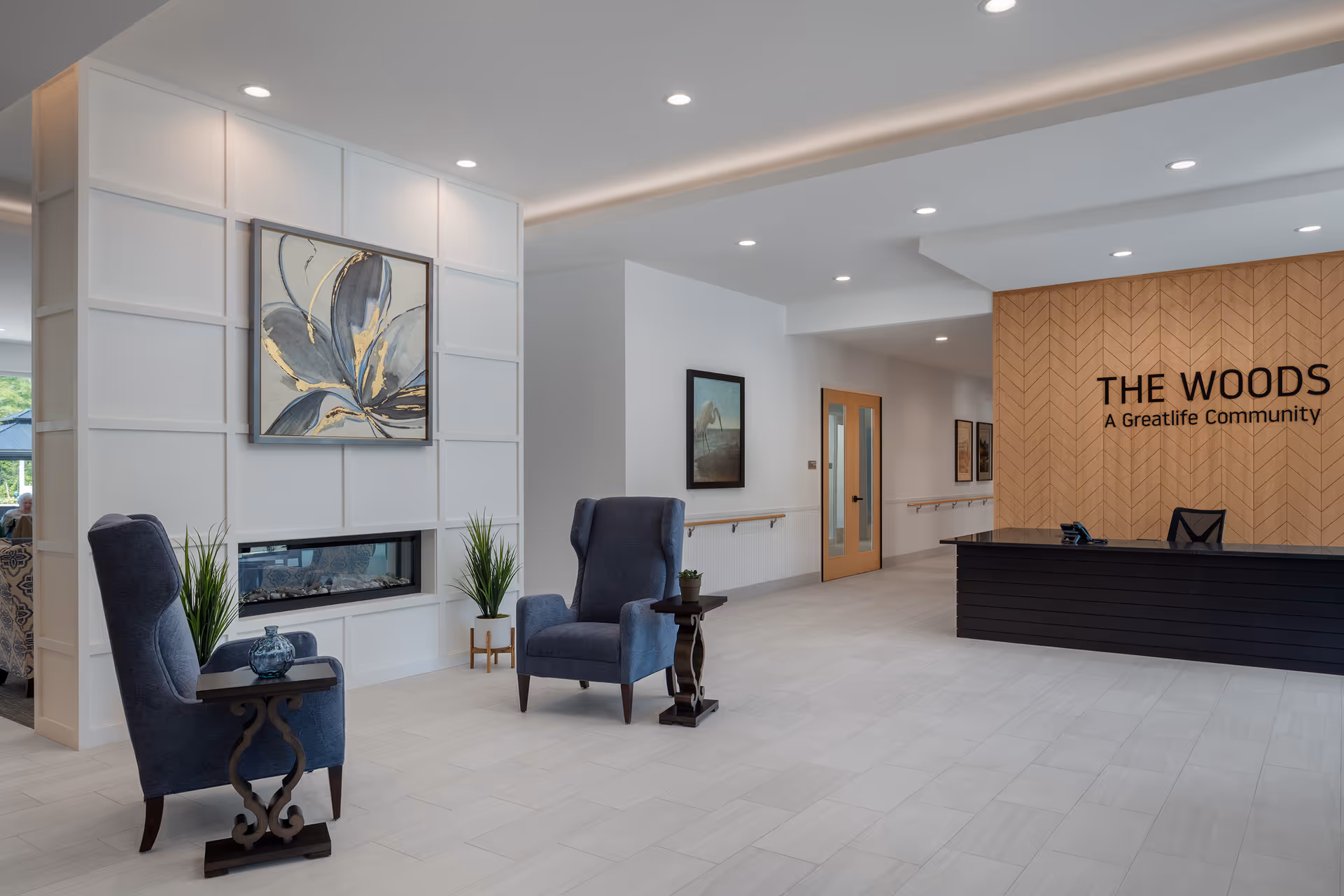A modern and spacious reception area of The Woods - A GreatLife Community featuring a black reception desk against a wood-paneled wall with the community name displayed. The room includes two blue armchairs with small side tables, a decorative fireplace, potted plants, and framed artwork on the walls. The floor is tiled in light gray, and the ceiling has recessed lighting.