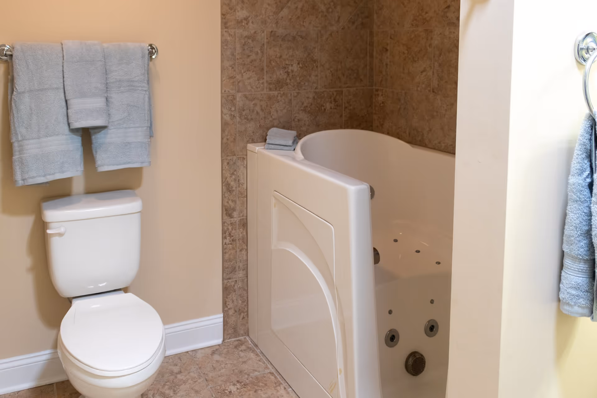 A bathroom featuring a white toilet with a towel rack above it holding three folded light blue towels. Next to the toilet is a walk-in bathtub with a door, surrounded by beige tiled walls and floor. A folded towel is placed on the edge of the bathtub.