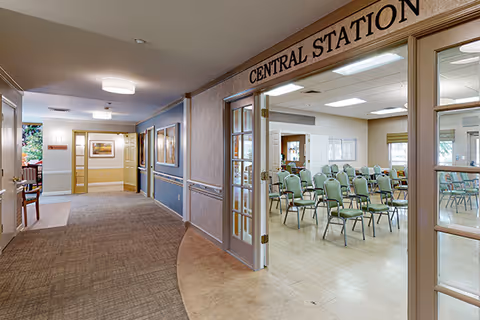 Interior view of a senior living facility hallway leading to a room labeled 'Central Station' with glass double doors. Inside the room, rows of green chairs are arranged facing a front area, suggesting a meeting or activity space. The hallway has carpeted flooring and walls decorated with framed pictures.