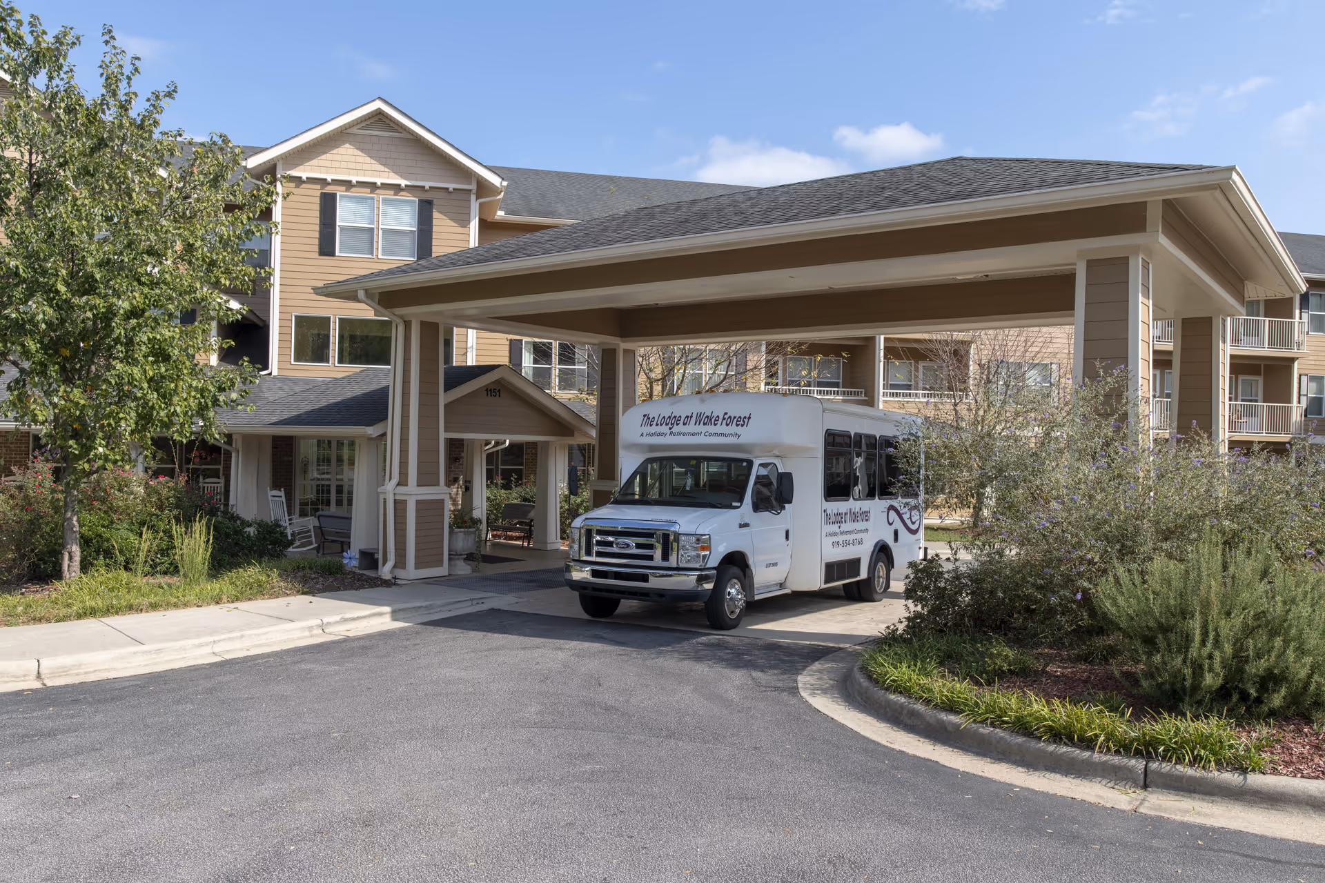 Exterior view of The Lodge at Wake Forest Gracious Retirement Living building with a covered entrance and a white shuttle bus parked underneath. The building is surrounded by greenery and trees under a partly cloudy sky.