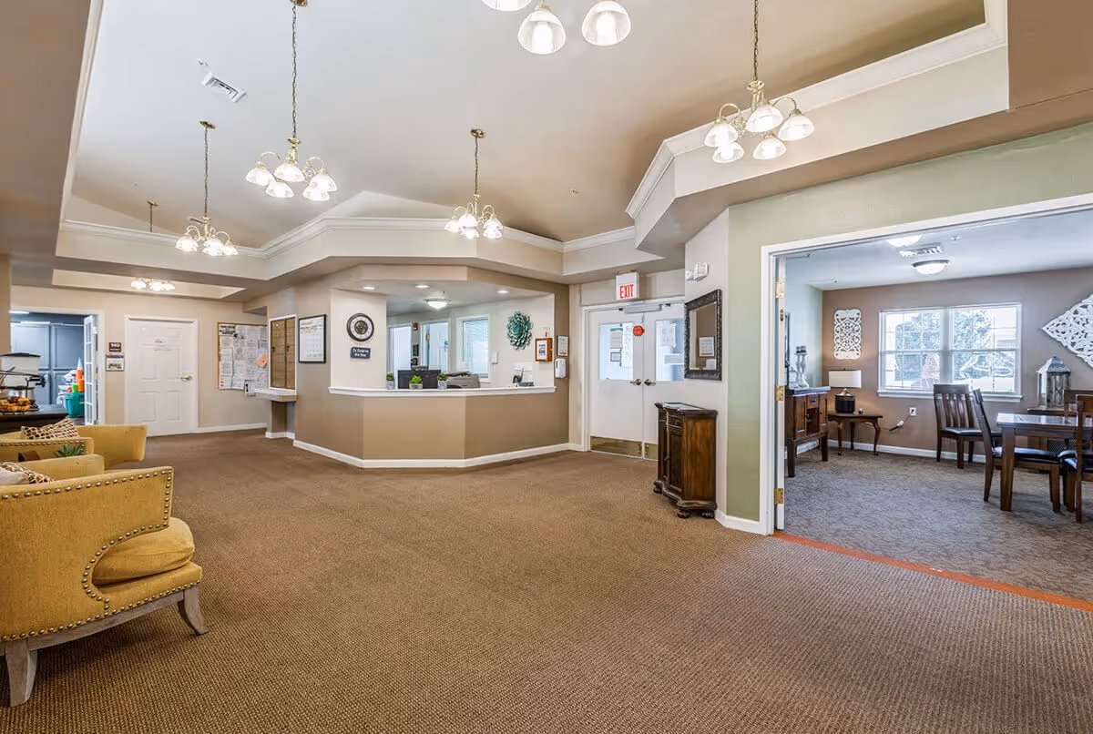 Interior view of a senior living facility reception area with a beige carpet, multiple ceiling light fixtures, a reception desk, and a seating area with yellow upholstered chairs. To the right, there is an open doorway leading to a room with a dining table and chairs, windows, and decorative wall art.