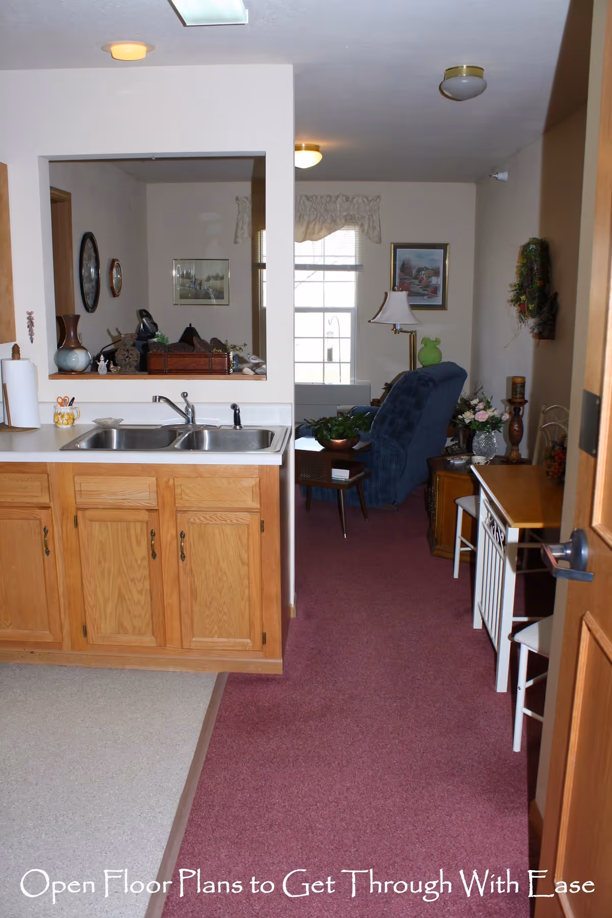 Kitchen sink and wooden cabinets open to a carpeted living room with a recliner and window.