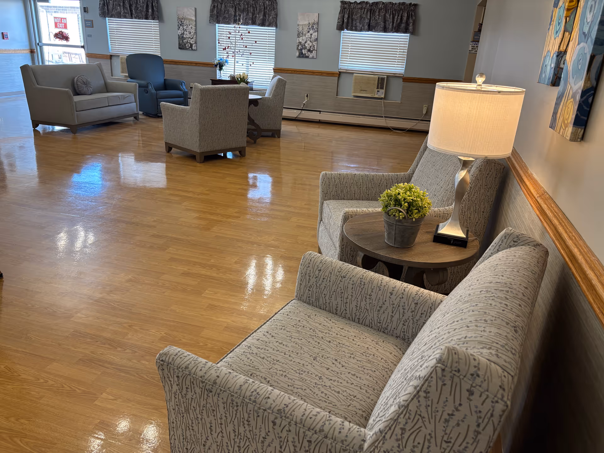 A spacious, well-lit common area with wooden flooring featuring multiple seating arrangements including patterned armchairs, a small round table with a lamp and a potted plant, a gray loveseat, and a blue armchair. The room has windows with blinds and valances, wall decorations, and an air conditioning unit mounted below one window.