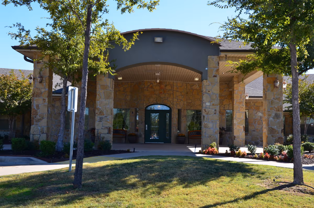 Front entrance of a nursing and rehabilitation facility with stone pillars, a covered entryway, glass double doors, and landscaped greenery including trees and shrubs.