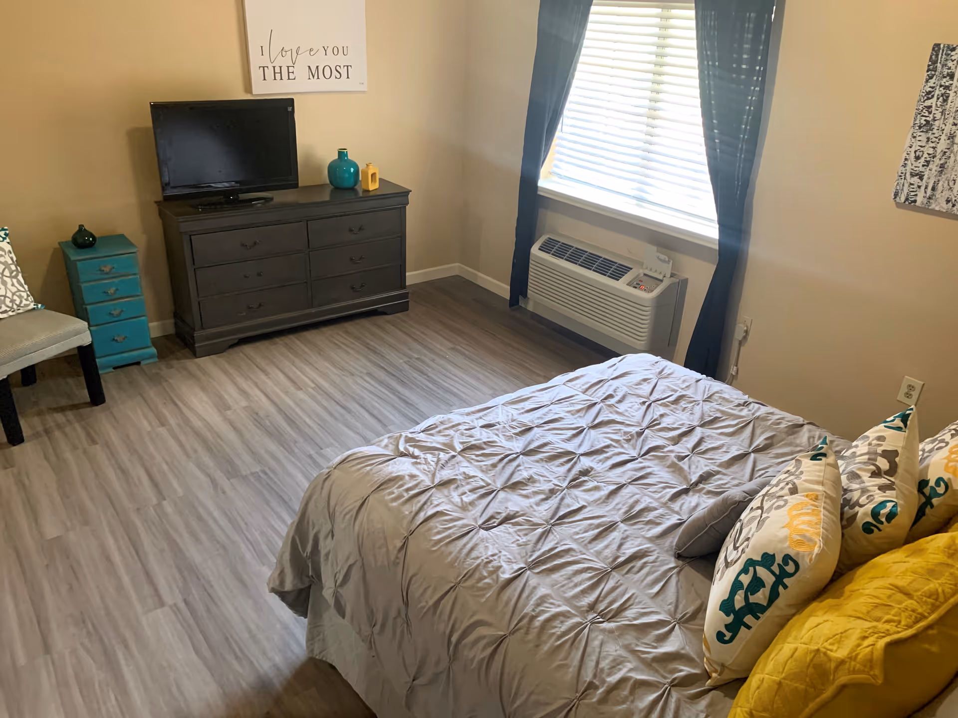 A cozy bedroom with a bed covered in a gray comforter and decorative pillows in yellow, white, and teal. There is a dark wooden dresser with a small flat-screen TV on top, along with two decorative vases. A teal-colored small drawer unit and a chair are placed to the left. A window with blinds and dark curtains is on the right wall above an air conditioning unit. A wall decoration above the dresser reads 'I love you THE MOST'.