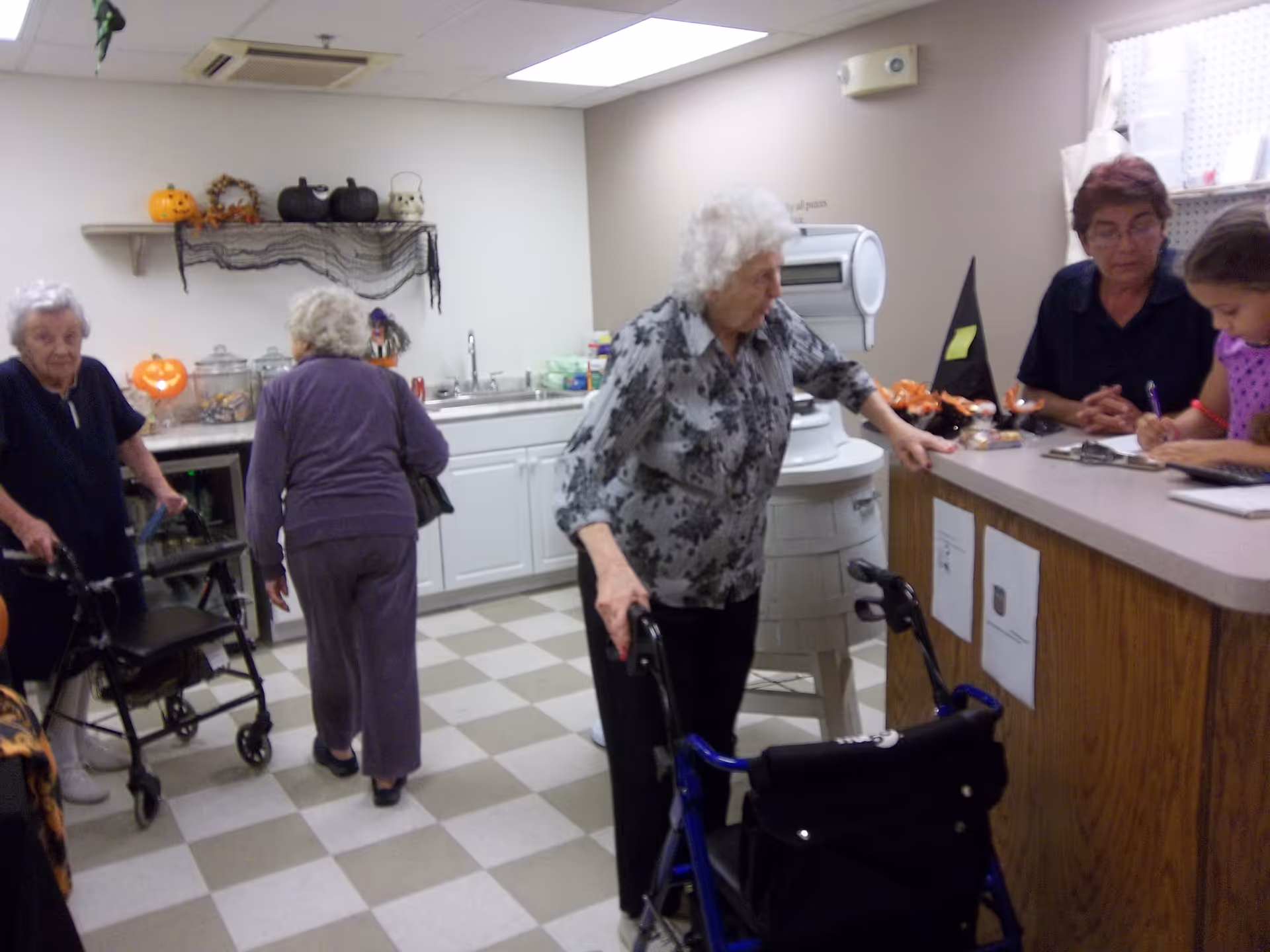 A group of elderly women in a room with a checkered floor and white cabinets. Two women are using walkers, one woman is walking away from the camera, and another woman is leaning on a counter where a staff member and a young girl are seated. The room is decorated with Halloween-themed items including pumpkins and a witch hat.
