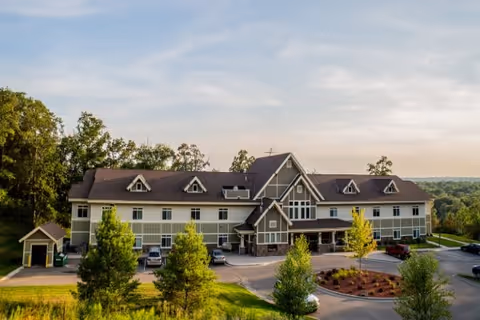 Exterior view of a large two-story assisted living facility building with a peaked roof and multiple dormer windows, surrounded by trees and greenery under a partly cloudy sky.