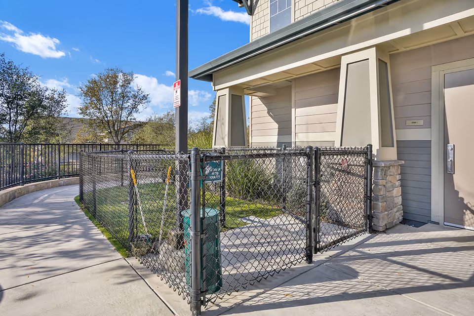 Outdoor fenced area with a small gate and a green pet waste disposal station, adjacent to a building with beige siding and stone accents under a clear blue sky.