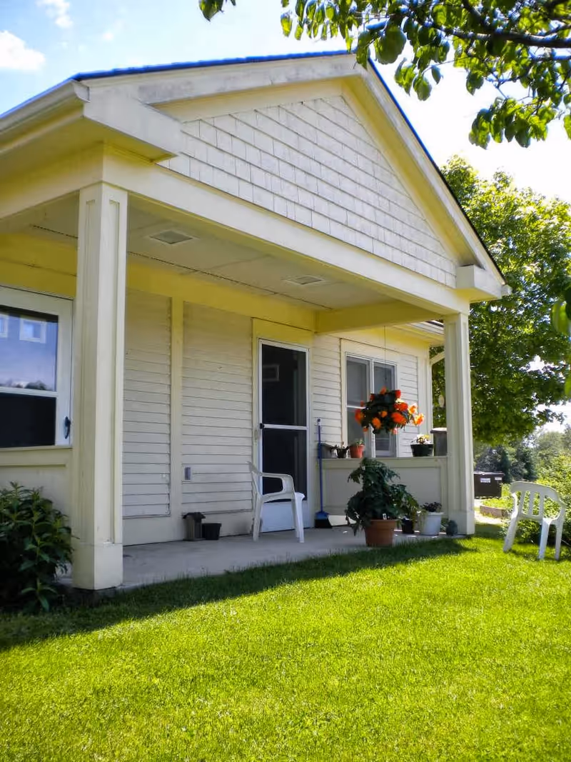 Front porch of a light-colored single-story building with a covered patio, plastic chairs, potted plants, and a green lawn.