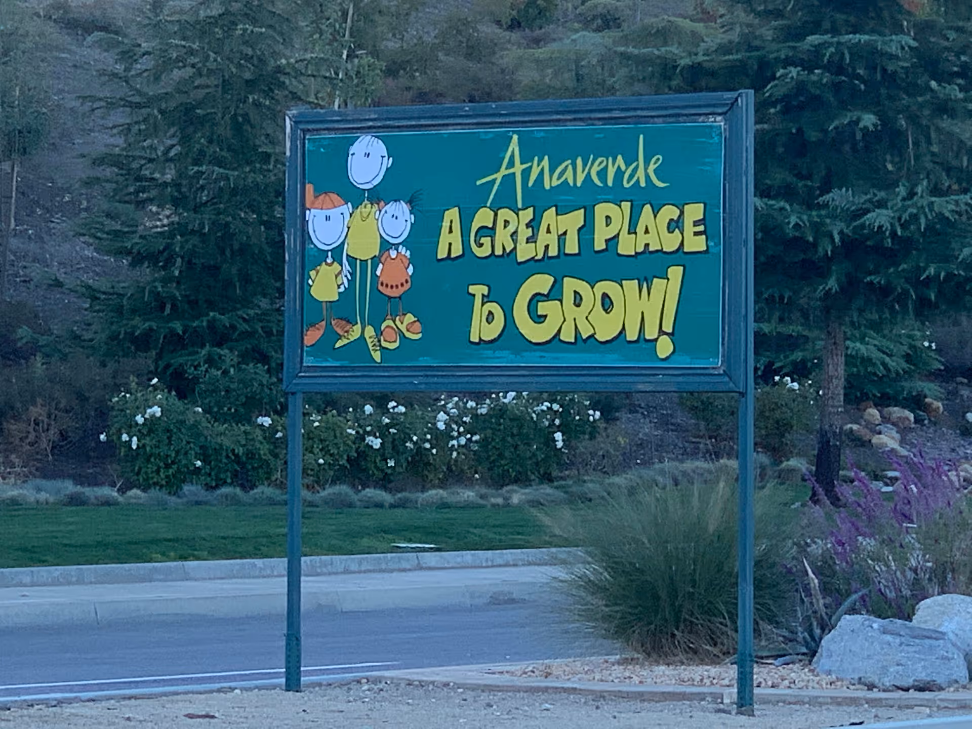 Outdoor signboard with cartoon drawings of three children and text that reads 'Anaverde A GREAT PLACE to GROW!' surrounded by trees, bushes, and a road.
