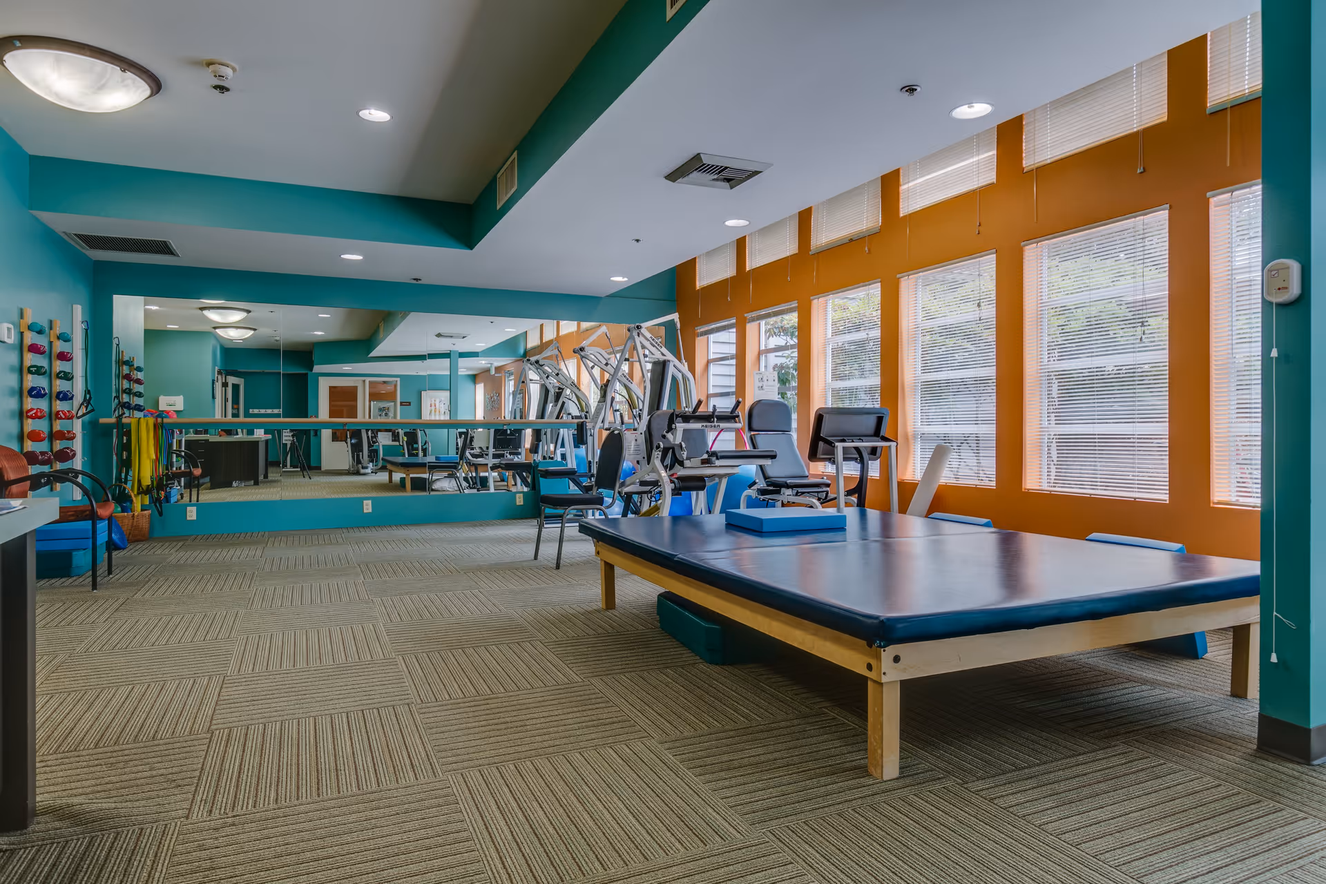 A spacious fitness or physical therapy room with exercise equipment including a large padded table, chairs, and various weights on racks. The room has teal and orange walls, large windows with blinds letting in natural light, and a large mirror covering one wall.