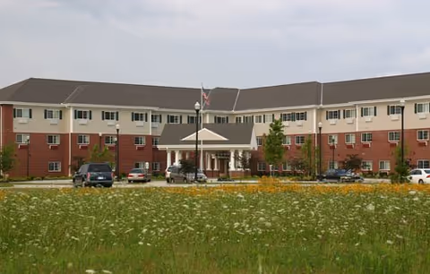 A large, multi wing senior living building with an entrance, parking, and a grassy field.