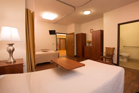 A senior living facility bedroom with two beds covered in white bedspreads, a wooden bedside table with a lamp, a wooden chair, and a dresser with a vase of flowers. There is a wall-mounted TV and an open door leading to a bathroom with a visible toilet.