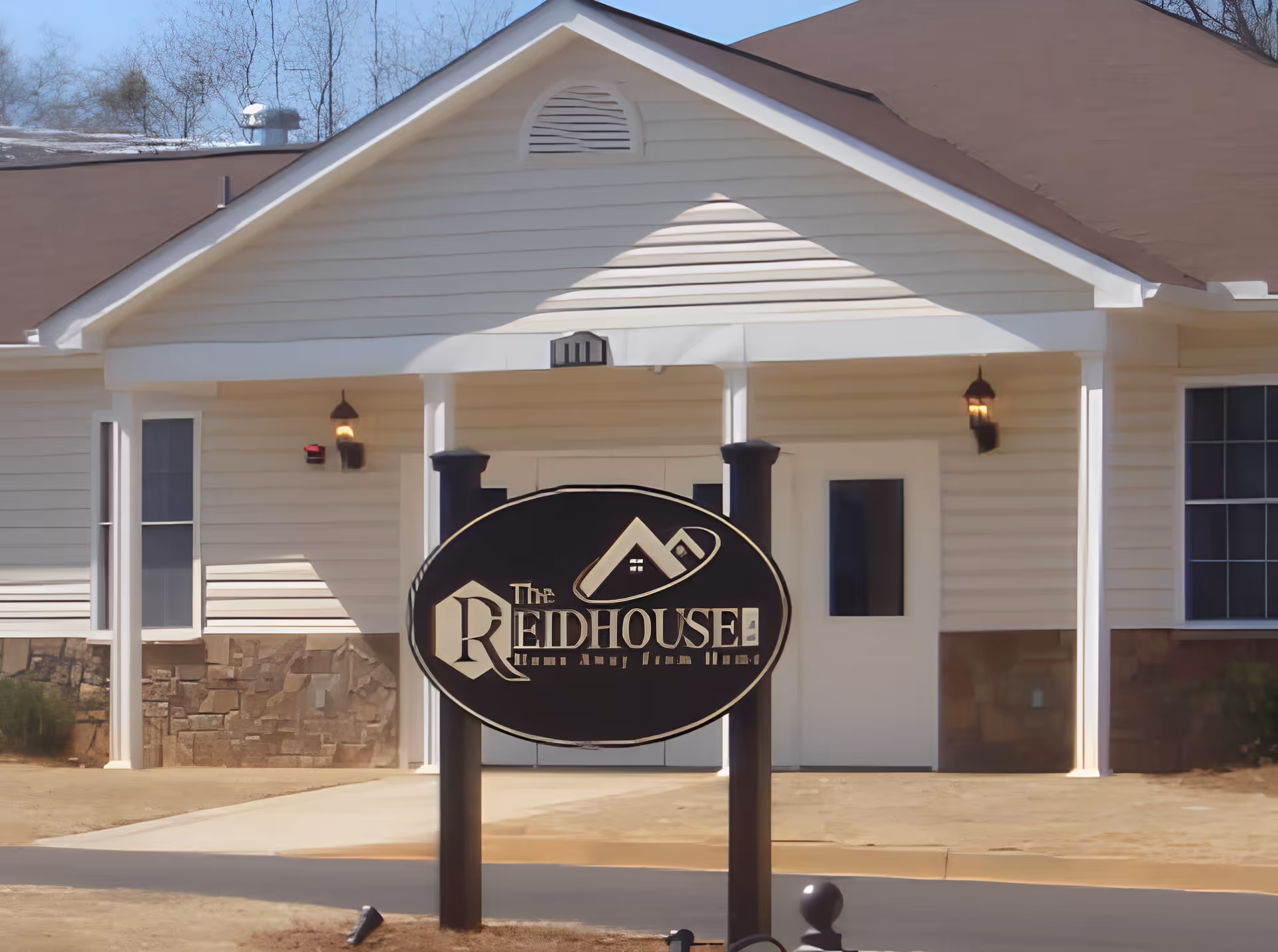 Exterior front view of a single-story building with beige siding and a brown roof. There is a large black oval sign in front with the text 'The Reid House' and a house logo. The building has two windows and a white door under a covered porch with two light fixtures.