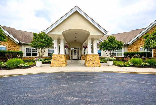 Front exterior view of a senior living facility with a covered entrance supported by white columns and stone bases, surrounded by well-maintained landscaping including green shrubs, trees, and flower pots.
