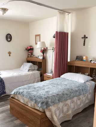 A simple, clean bedroom with two single beds separated by a curtain. Each bed has a wooden headboard with personal items and decorations, including a lamp, flowers, and framed pictures. The room has light-colored walls with religious crosses hanging on them and a wooden floor.