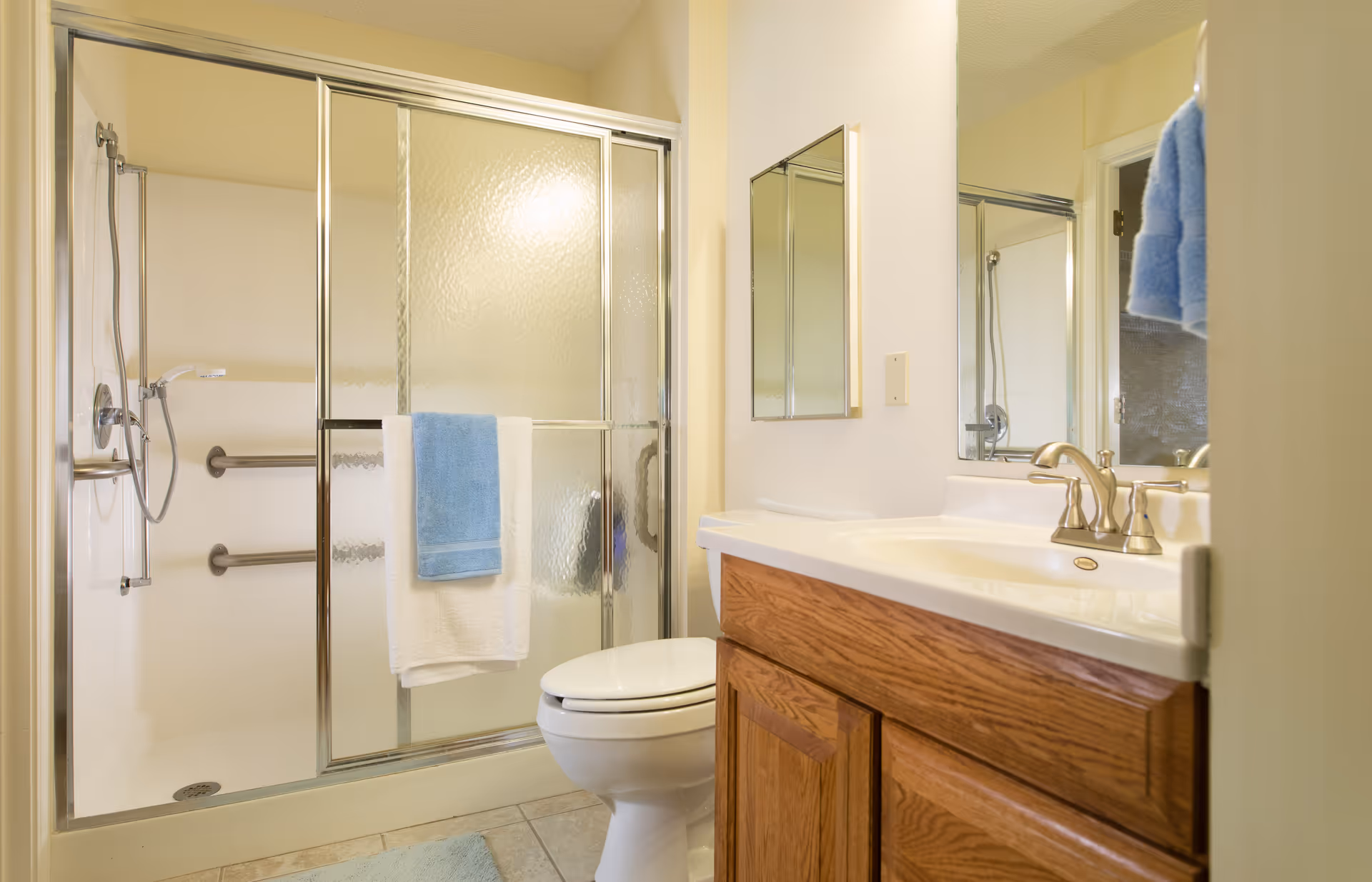 A clean bathroom featuring a shower with sliding glass doors and grab bars, a white toilet, a wooden vanity with a white sink and silver faucet, a mirror above the sink, and blue and white towels hanging on the shower door and wall.