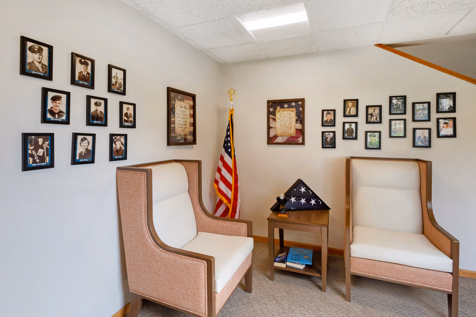 A small seating area with two beige and brown armchairs facing each other with a wooden side table in between. On the table is a folded American flag in a display case and a small eagle figurine. The walls are decorated with framed black and white photographs of military personnel and two framed documents with American flag backgrounds. An American flag stands in the corner of the room.