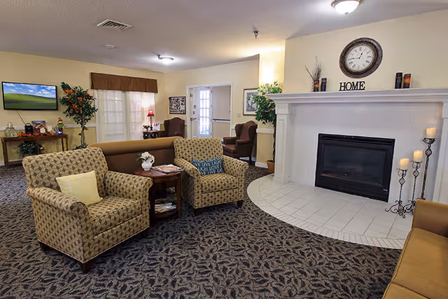 A cozy living room area in a senior living facility with patterned armchairs, a small wooden side table with a flower vase and magazines, a large white fireplace with candles and a clock above it, and a TV on a stand in the background. The room has carpeted flooring and soft yellow walls with framed pictures and plants.