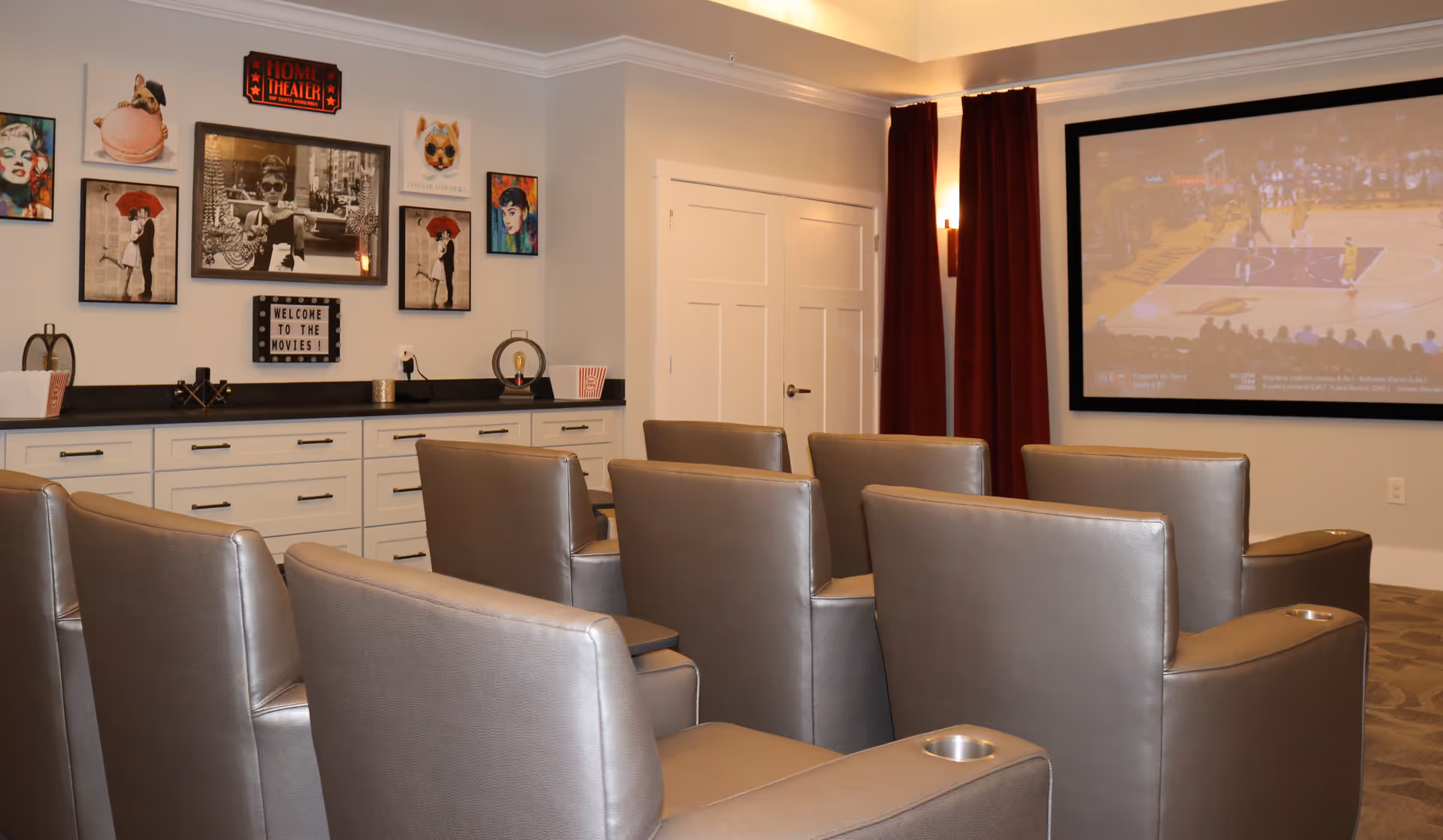 A cozy home theater room with six gray leather recliner chairs arranged in two rows facing a large screen showing a basketball game. The walls are decorated with various framed pictures and a sign that reads 'WELCOME TO THE MOVIES!'. There is a long white cabinet with black countertop along the left wall, and red curtains hang beside the screen.