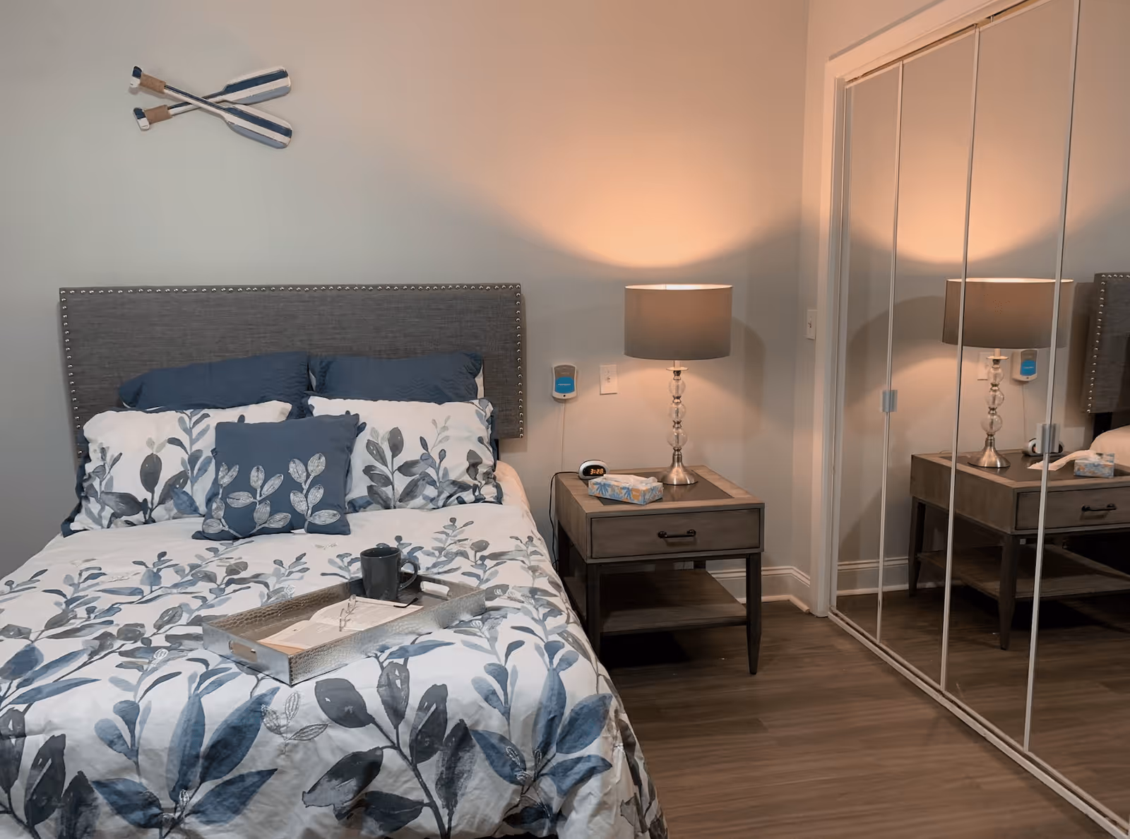 A cozy bedroom with a bed featuring a gray upholstered headboard and blue and white floral bedding. A wooden nightstand with a drawer and an open shelf sits beside the bed, holding a lamp with a gray shade, a tissue box, and a small clock. Two decorative oars are mounted on the wall above the bed. A mirrored closet door reflects the nightstand and lamp.