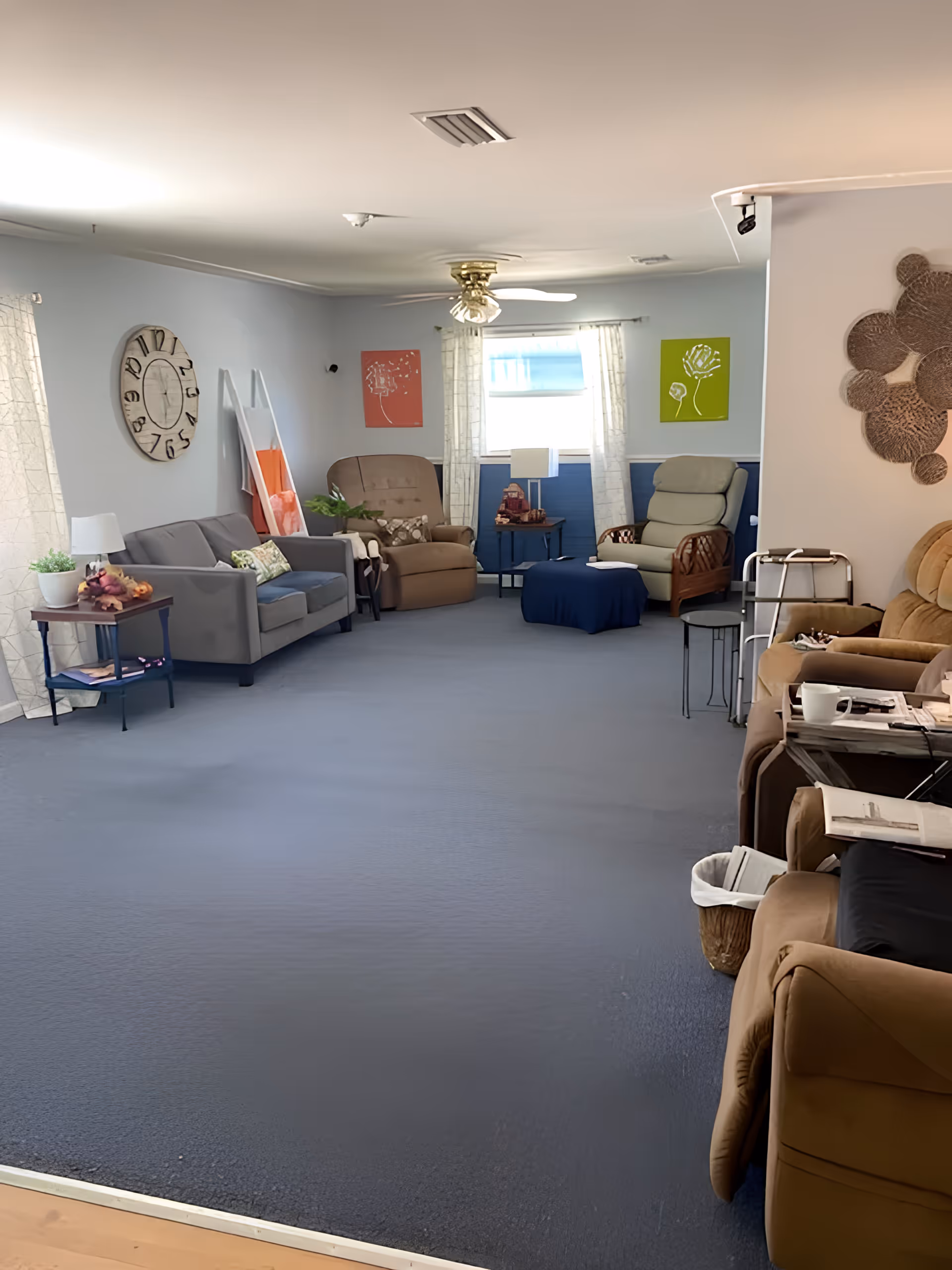 Bright communal living room with sofas and recliner chairs arranged around side tables, wall art, and a ceiling fan.