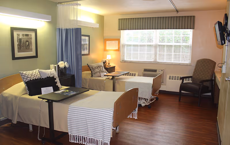 A nursing facility room with two hospital-style beds separated by a blue privacy curtain. Each bed has a beige blanket, pillows, and a small overbed table. There is a nightstand with a lamp and flowers between the beds. A large window with blinds lets in natural light. A cushioned armchair is placed near the window, and a wall-mounted TV is visible on the right side. The room has wooden flooring and soft lighting.