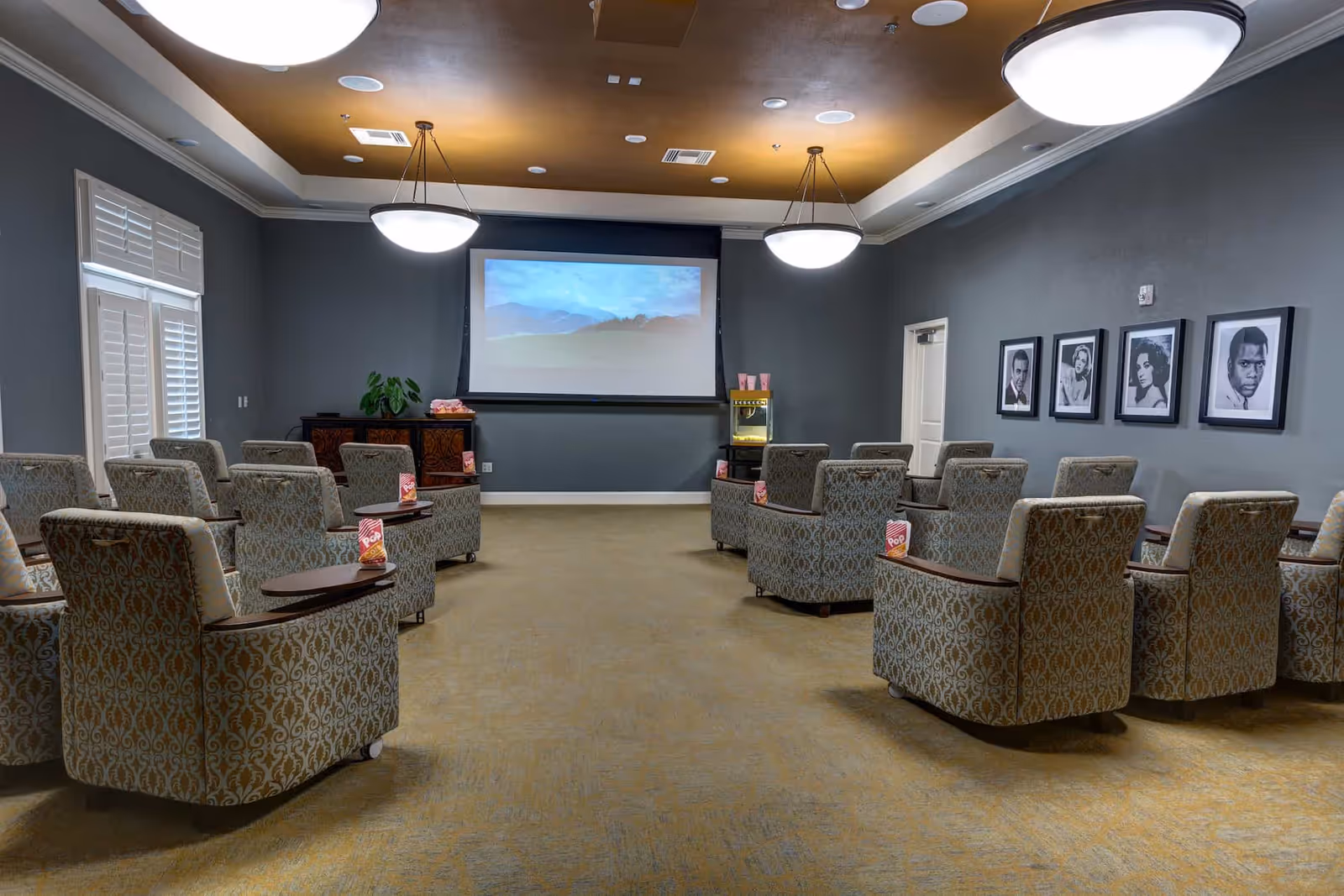 A small theater room with patterned armchairs arranged in rows facing a large projection screen. Each chair has a small wooden tray with a popcorn container. The room has gray walls, a gold ceiling with two large round light fixtures, and framed black and white portraits on one wall. There is a popcorn machine and a cabinet at the front of the room.