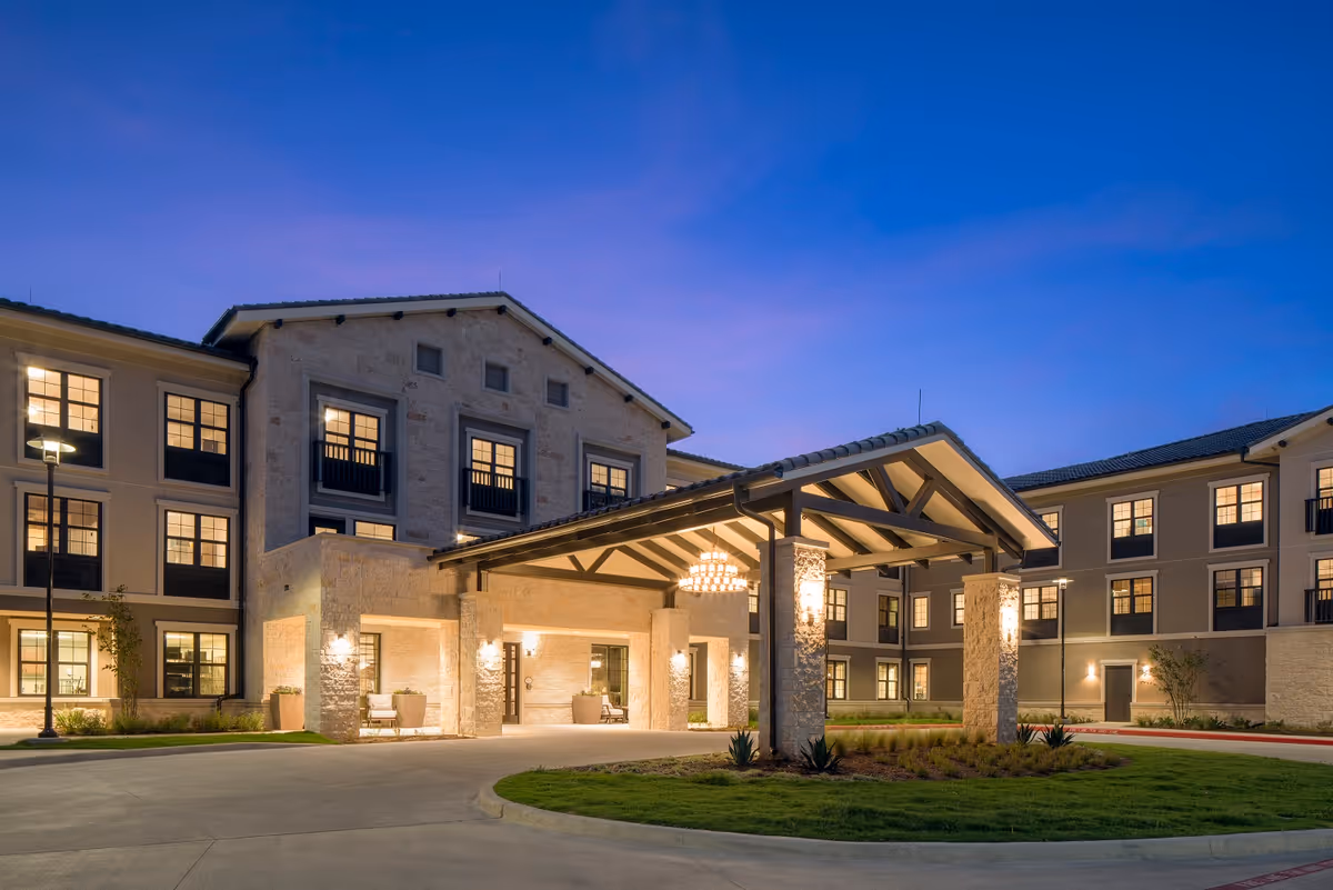Front entrance of a three-story senior living building with a covered porte-cochere and warm exterior lighting at dusk.