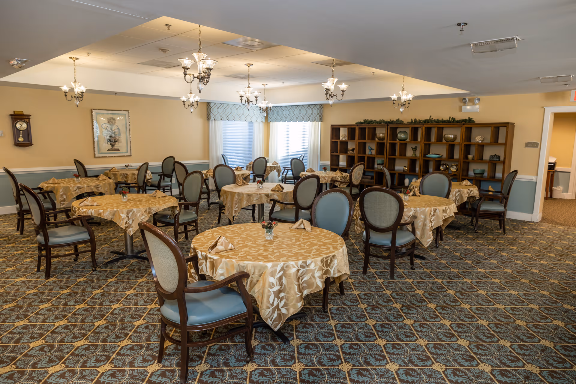 A dining room with multiple round tables covered with beige patterned tablecloths, each set with folded napkins and small flower arrangements. The room has upholstered chairs with wooden frames, a patterned carpet, chandeliers hanging from the ceiling, a wall clock, framed artwork, and a large wooden shelving unit with decorative items. Windows with curtains allow natural light into the room.