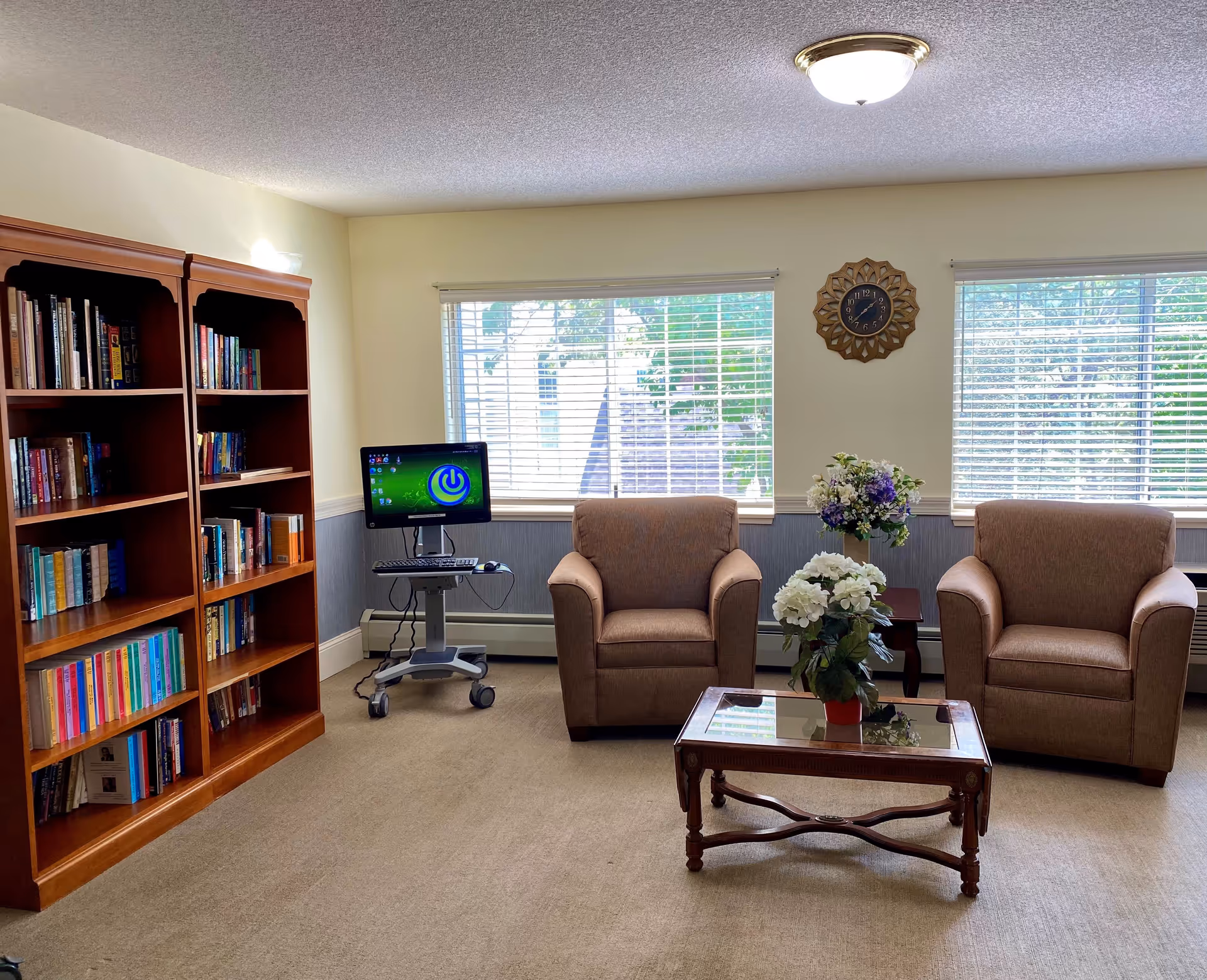 Cozy common room with two armchairs, a glass-top coffee table with flowers, a bookshelf, and a computer workstation in front of windows.