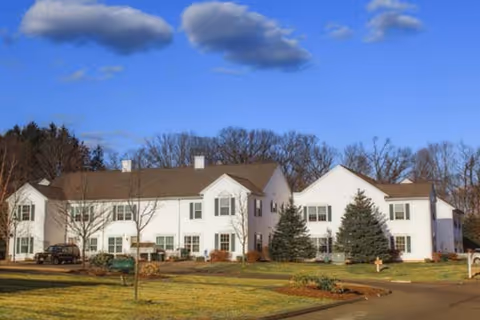 Exterior view of a large white two-story building with multiple windows and a brown roof, surrounded by a lawn with some trees and shrubs under a blue sky with scattered clouds.