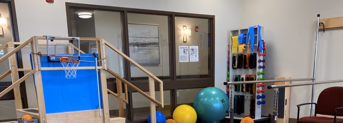 Indoor therapy or exercise room with wooden stairs and a small basketball hoop, various exercise balls, resistance bands, dumbbells, and a chair. The room has large windows and neutral-colored walls.