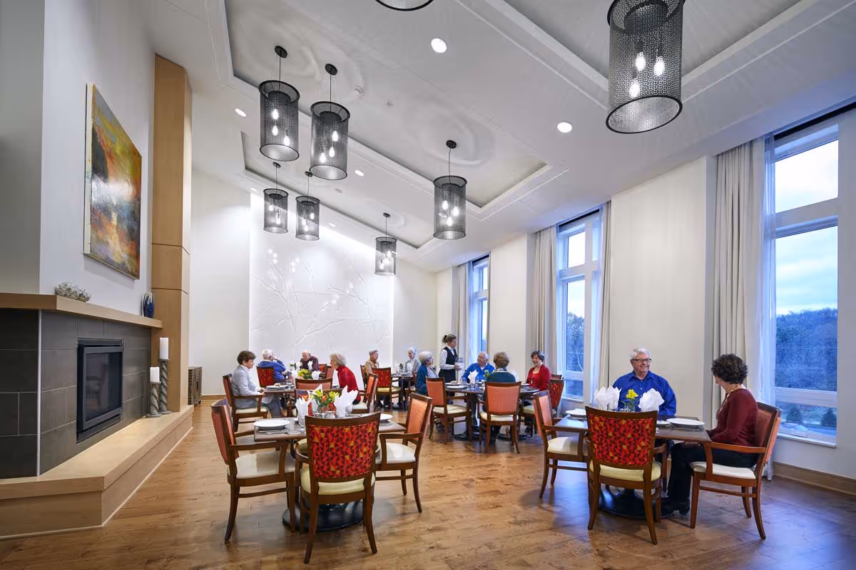 Seniors seated at tables in a bright, modern communal dining room with large windows and hanging pendant lights.