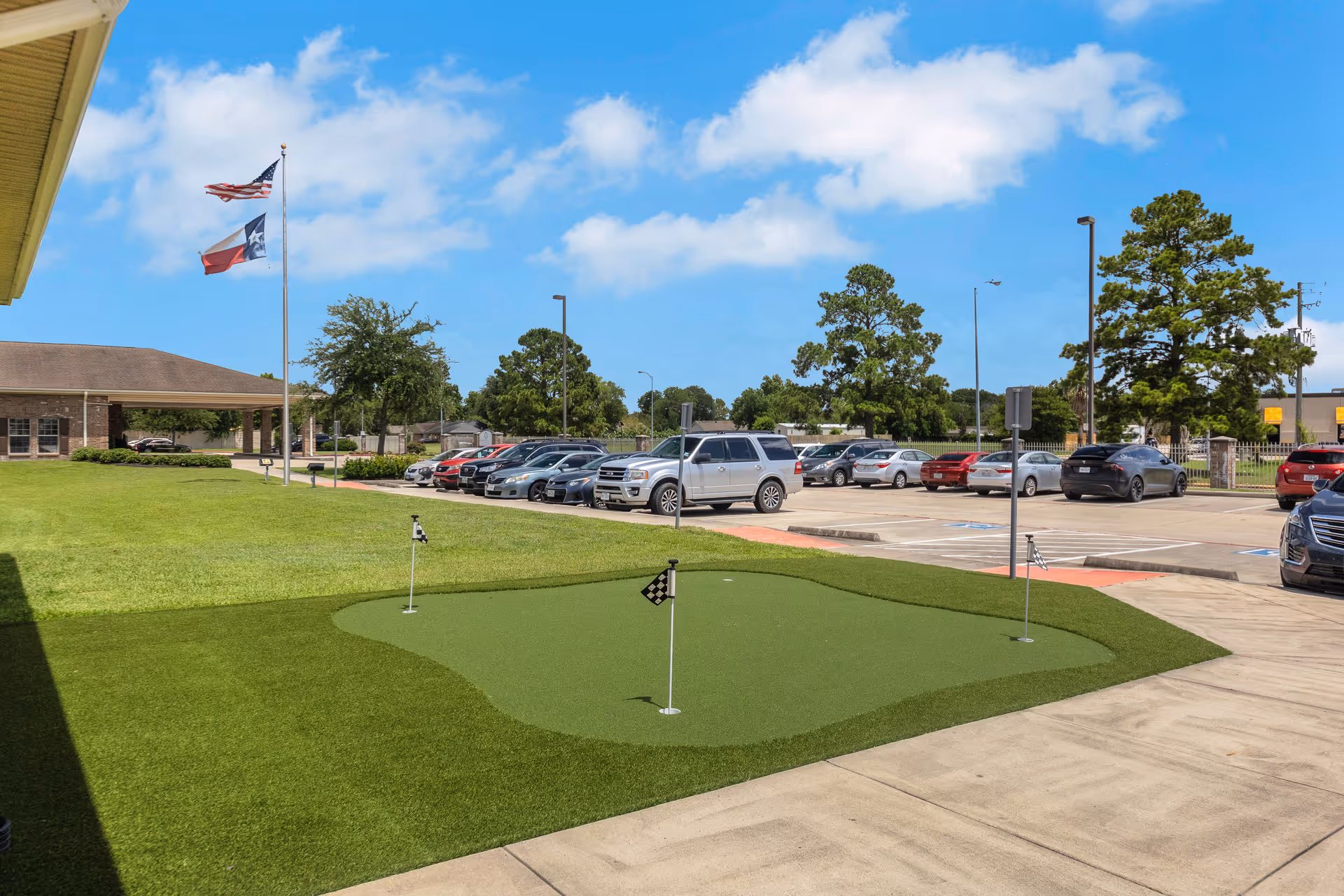 Front exterior of a senior living facility with a small putting green, parking lot, flags, and the building entrance under a blue sky.