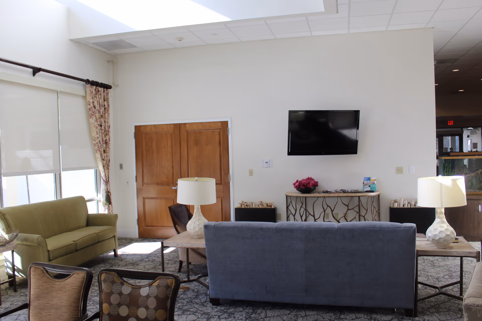 A bright living room area with a green sofa on the left, a blue sofa facing away from the camera, and a wooden door in the background. A flat-screen TV is mounted on the white wall above a decorative console table with a flower arrangement. Two table lamps with white shades are placed on side tables beside the sofas. Large windows with floral curtains and roller shades allow natural light to fill the room.