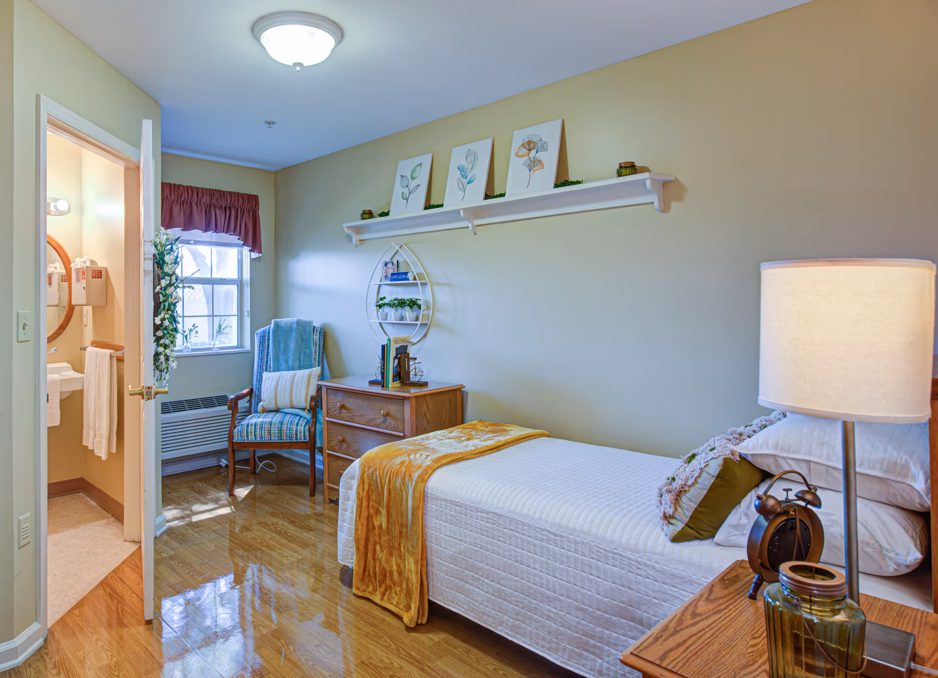 A cozy bedroom in a senior living facility with a single bed covered in a white quilt and a mustard yellow throw blanket. There is a wooden nightstand with a lamp and an alarm clock beside the bed. A wooden dresser with books and decorative items sits against the wall. Above the dresser, a white shelf holds three framed botanical prints and small plants. A comfortable armchair with a cushion and a blue throw blanket is placed near a window with a maroon valance. The door to a small bathroom is open, showing a sink, mirror, and towel rack.