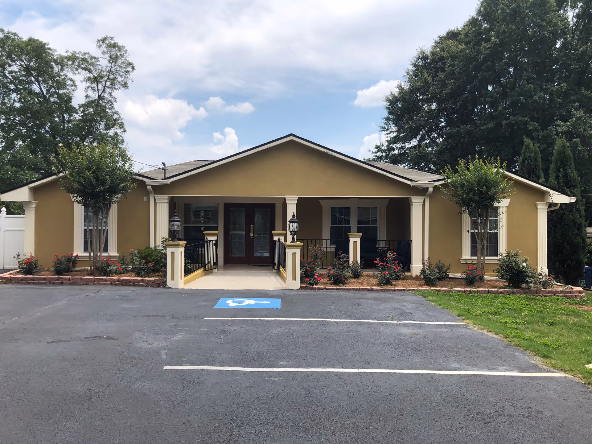 Front exterior view of a single-story building with a beige facade, white columns, and a covered entrance with double doors. There are two small trees and flower beds with red flowers on either side of the entrance. A parking lot with a handicapped parking space is visible in front of the building under a partly cloudy sky.