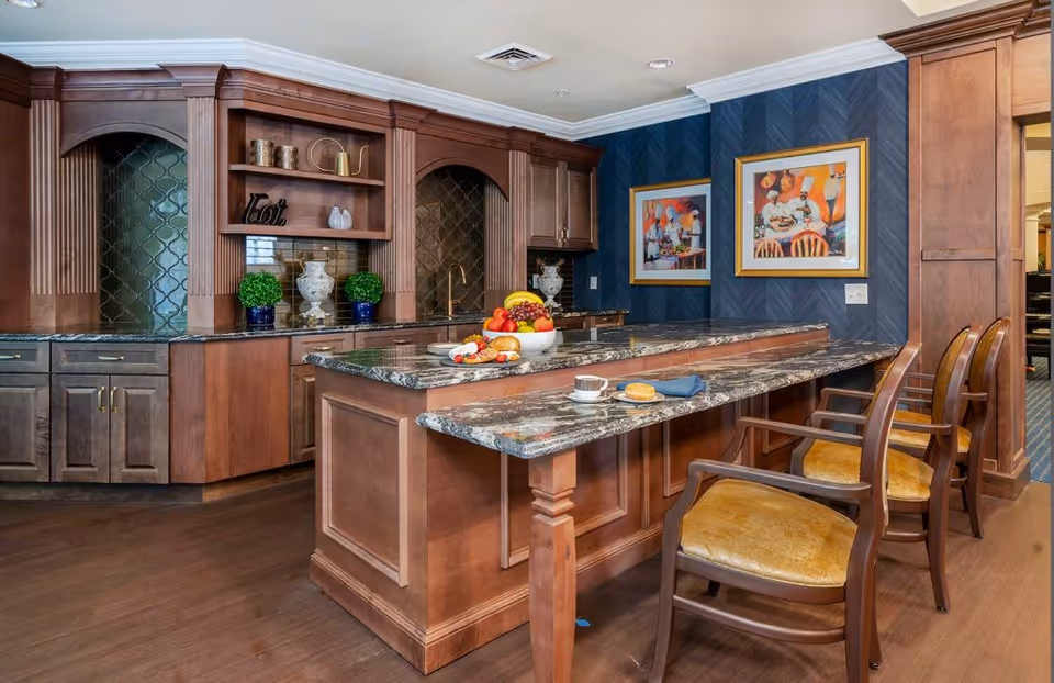A kitchen area with wooden cabinetry and a granite countertop island. The island has a raised bar section with three wooden chairs with yellow cushions. On the countertop, there is a bowl of assorted fruits, a plate with a croissant, a cup of coffee, and a blue napkin. The background features dark blue patterned wallpaper with two framed colorful paintings. The floor is wooden.