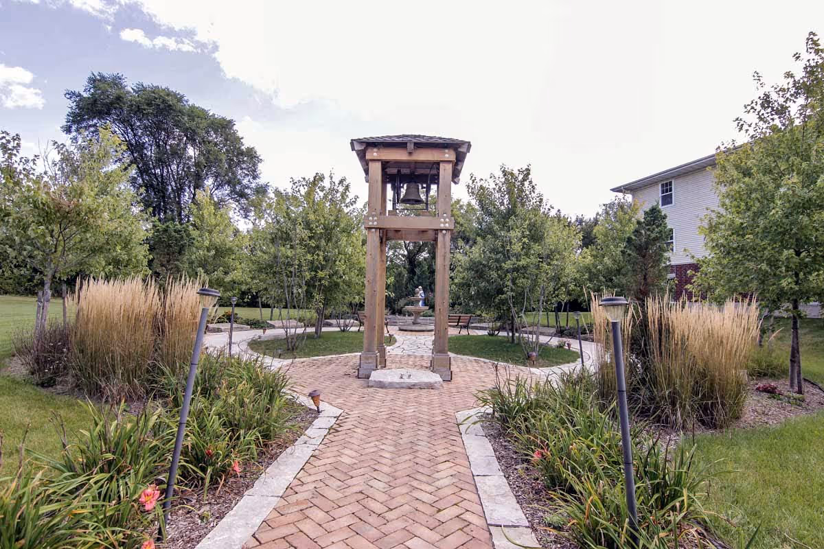 A landscaped outdoor garden area with a brick pathway leading to a wooden bell tower structure. The pathway is bordered by plants and small trees, with benches and a fountain visible in the background. A building is partially visible on the right side.
