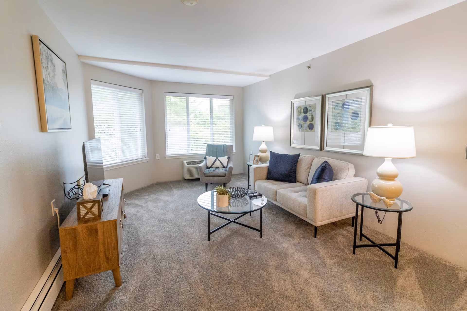 A bright and cozy living room with a beige sofa adorned with dark blue pillows, a glass coffee table with decorative items, a side table with a lamp, a gray armchair near large windows with blinds, and a wooden TV stand with a television and decorative items.