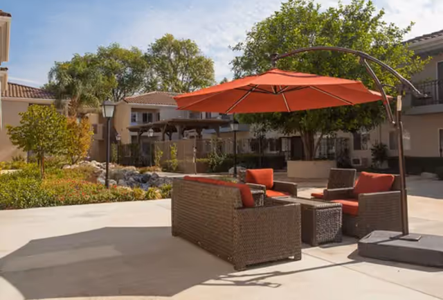 Outdoor patio area at Walnut Valley Senior Living featuring wicker seating with red cushions and a large red umbrella, surrounded by trees and residential buildings under a clear sky.