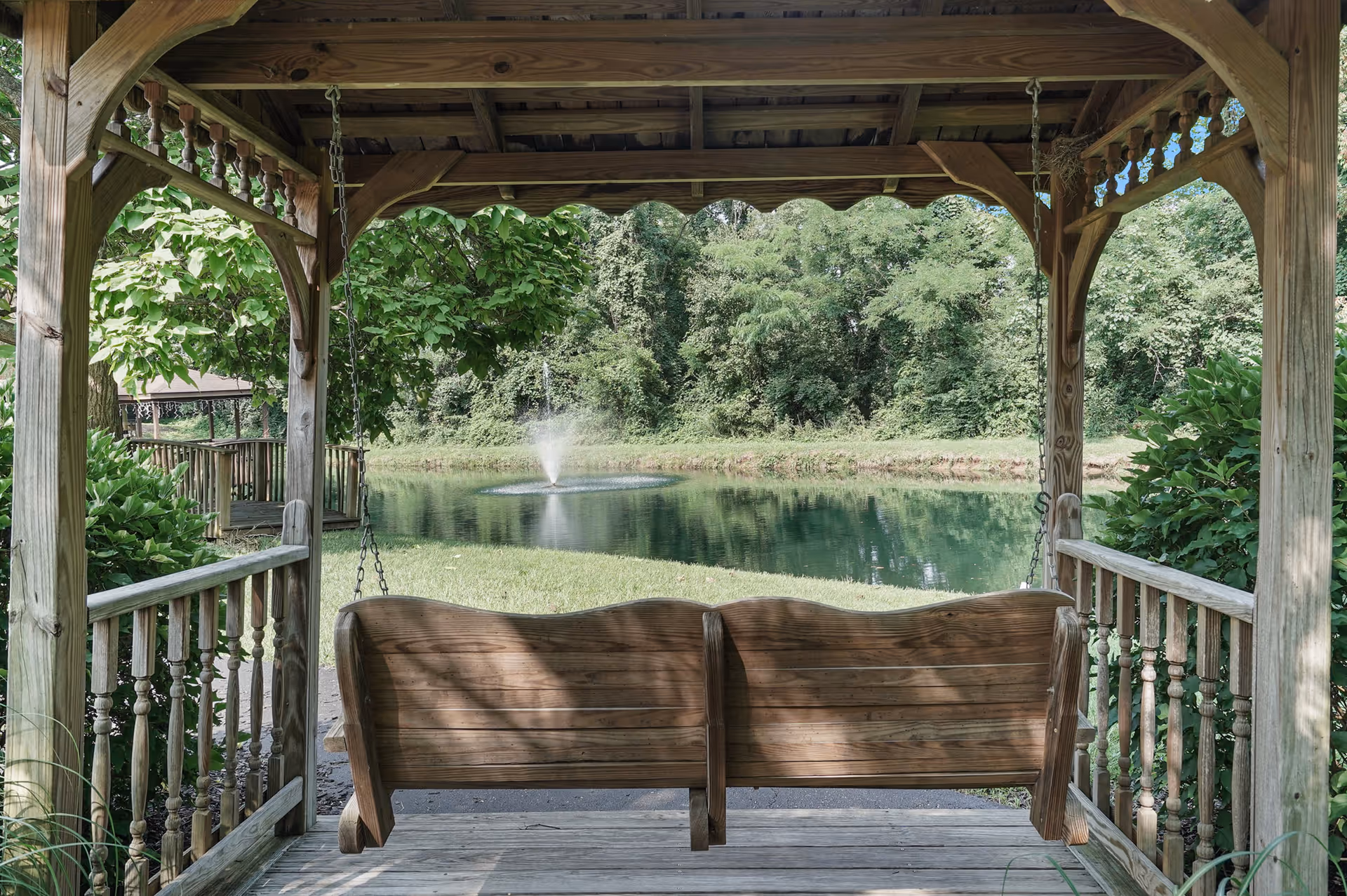 A wooden porch swing hanging under a wooden gazebo overlooking a small pond with a water fountain in the middle, surrounded by green trees and bushes.