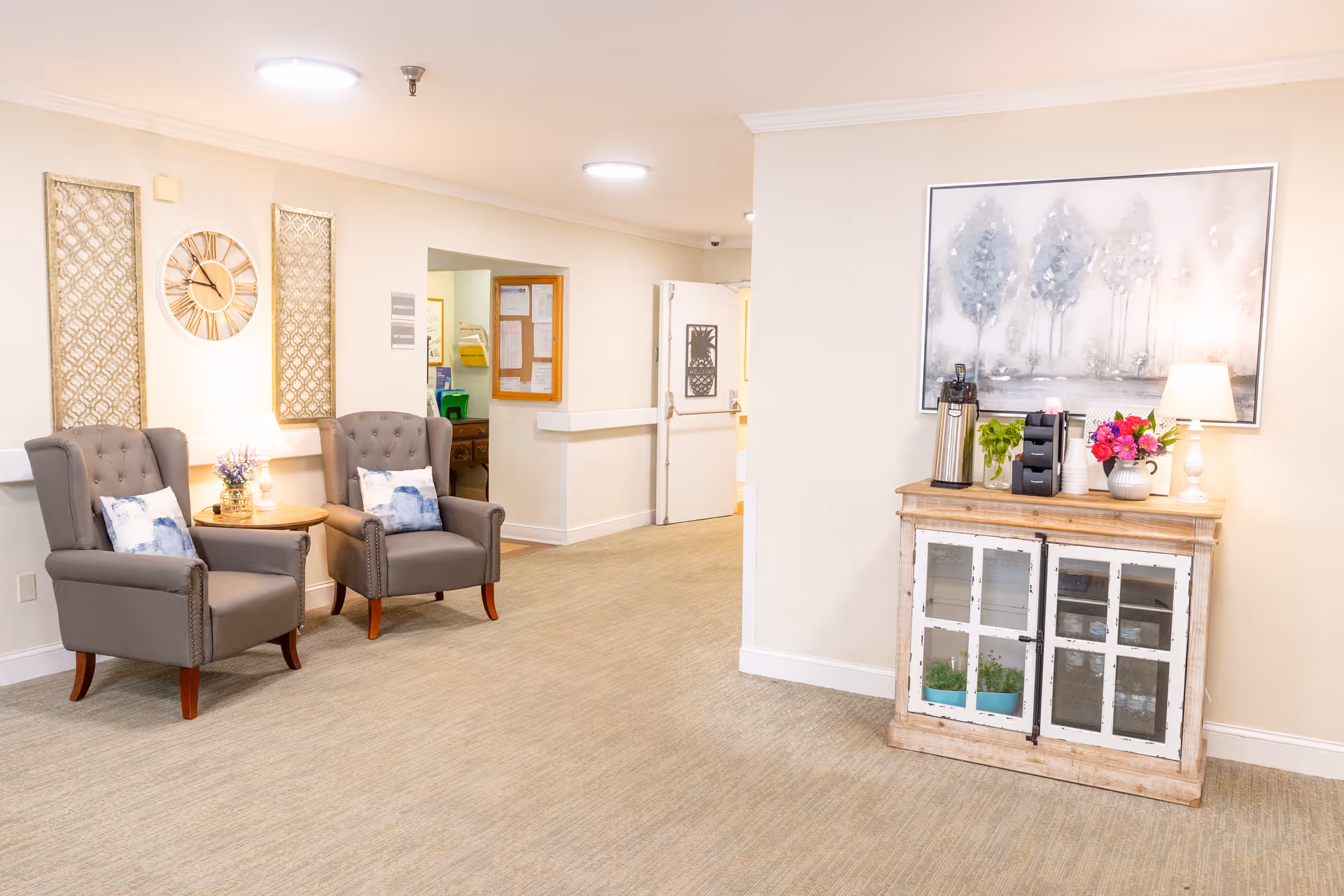 A bright and cozy interior hallway area in a nursing home featuring two gray upholstered armchairs with decorative pillows, a small round wooden table with a lamp and flowers, a wooden cabinet with glass doors holding plants and a coffee station on top, a large framed artwork of trees on the wall, and a door leading to another room.