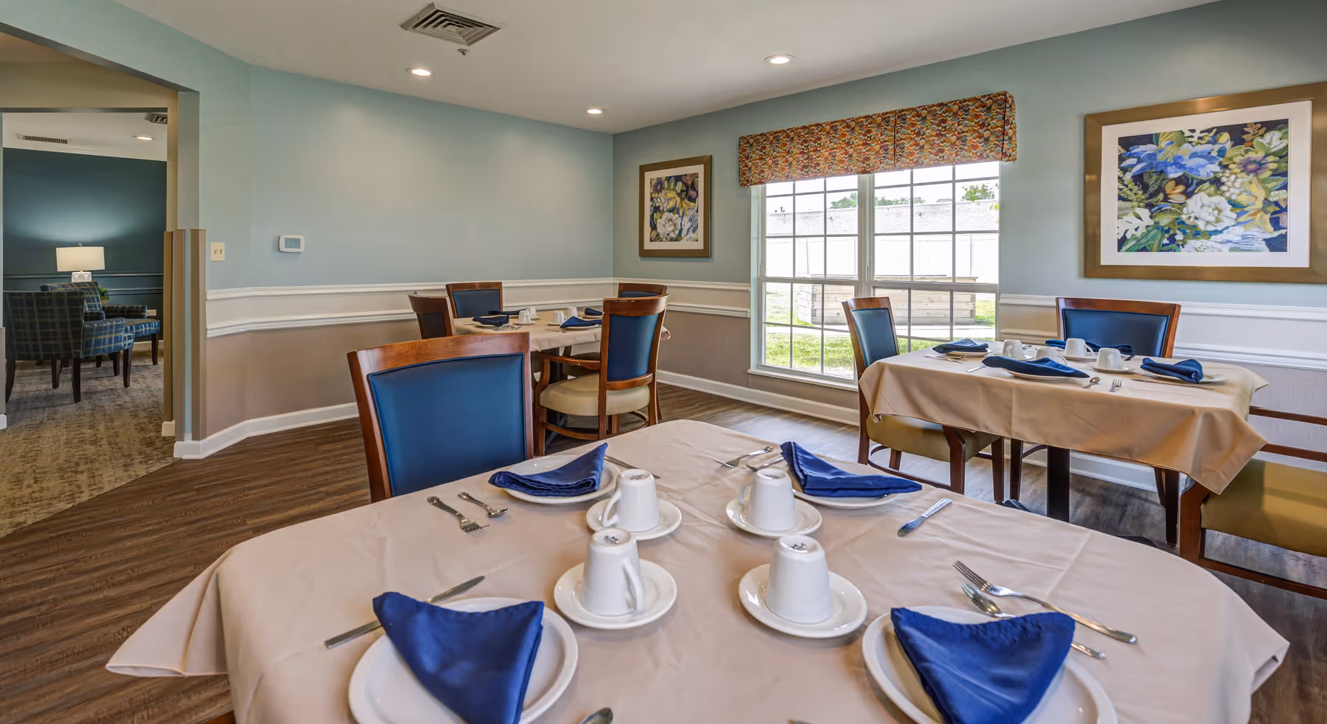 A dining room in a senior living facility with tables covered in beige tablecloths, each set with white plates, cups, silverware, and blue cloth napkins. The room has light blue and beige walls with white trim, two framed floral paintings, a large window with a patterned valance, and wooden chairs with blue cushions. In the background, there is an adjacent room with blue upholstered chairs and a lamp.