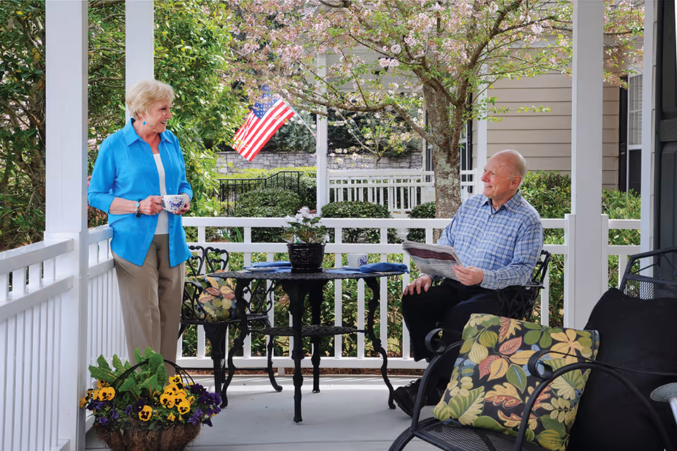 An elderly woman and man are enjoying a conversation on a porch. The woman is standing holding a cup, wearing a blue shirt and beige pants. The man is sitting on a chair holding a newspaper, wearing a blue checkered shirt and black pants. The porch is decorated with a small black table, chairs with floral cushions, a flower pot, and a basket of flowers. There is an American flag and blooming trees in the background.