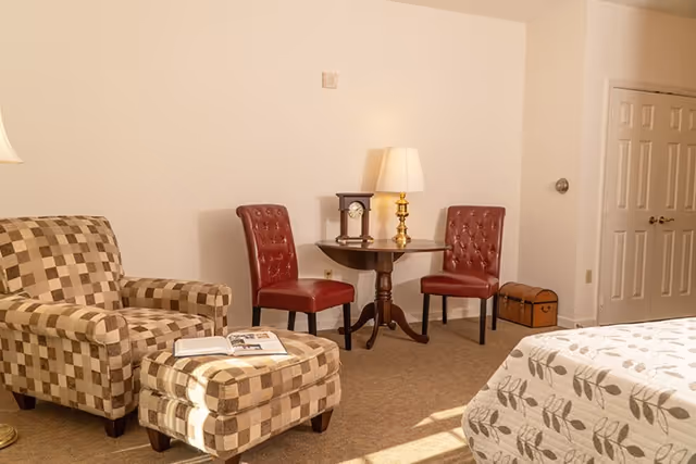 Cozy assisted living bedroom with a patterned armchair and ottoman, two red chairs at a small table with a lamp, and part of a bed visible.