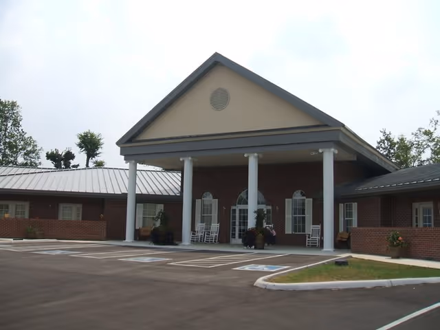 Front exterior view of Trousdale Senior Living Center showing a brick building with a covered entrance supported by white columns, several windows, rocking chairs on the porch, and a parking lot with marked handicap spaces in front.