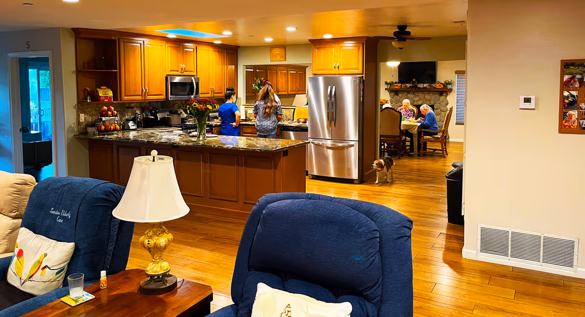 Interior view of a senior living facility showing a living room area with blue recliners and a wooden side table with a lamp. In the background, there is a kitchen with wooden cabinets, a granite countertop island, and two people standing near the counter. Further back, two elderly individuals are seated at a dining table near a stone fireplace, and a small dog is walking on the wooden floor.