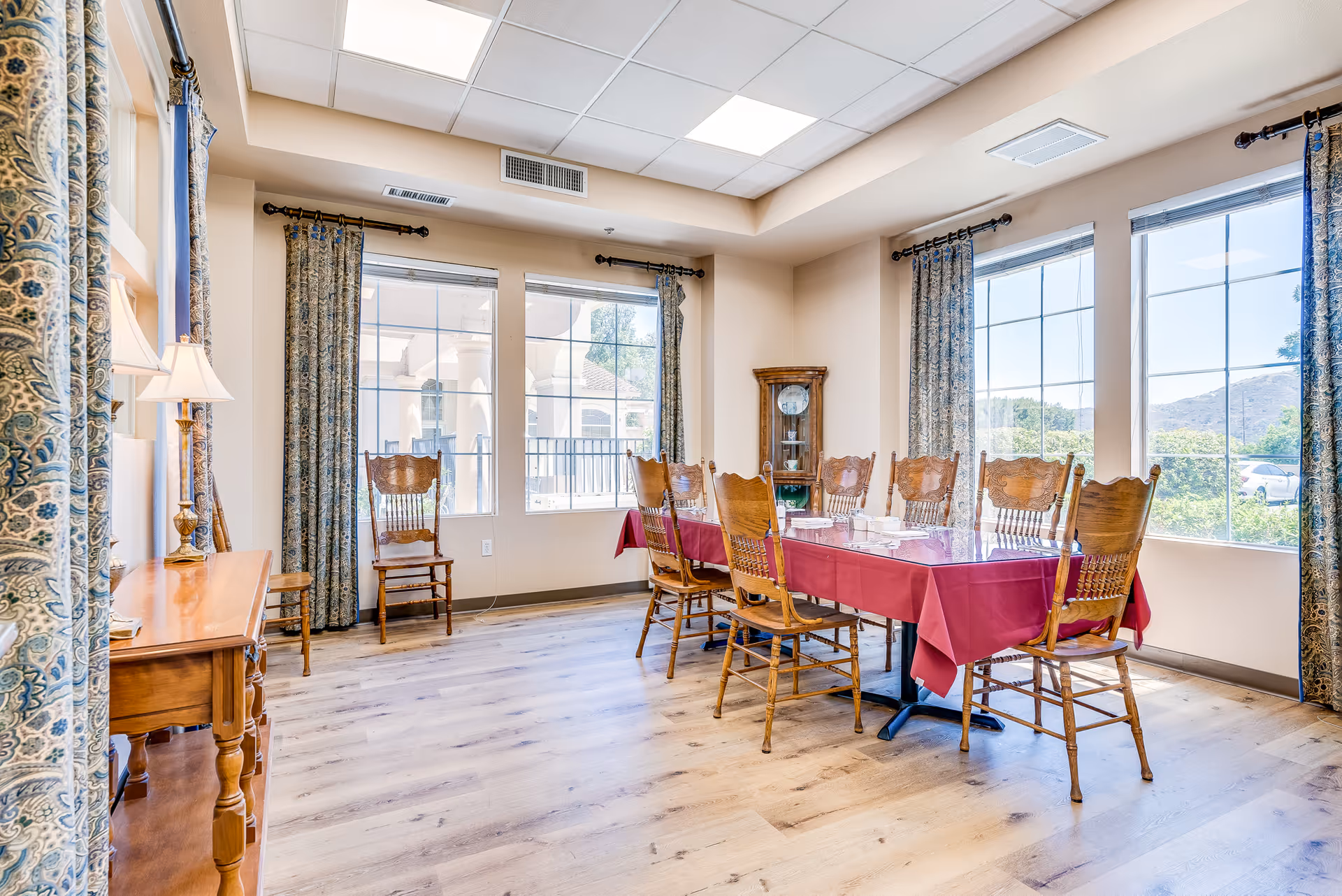 Bright dining room with a long table covered in a red tablecloth surrounded by wooden chairs and large windows.